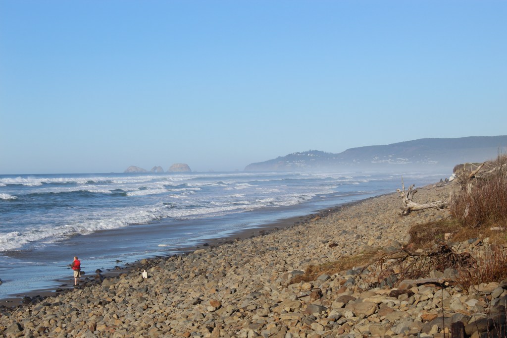 Rocky beach and the crashing waves of the Pacific Coast. A man with a white beard and red vest is on the shoreline fishing. In the far background are a town on a coastal hillside and rock formations.
